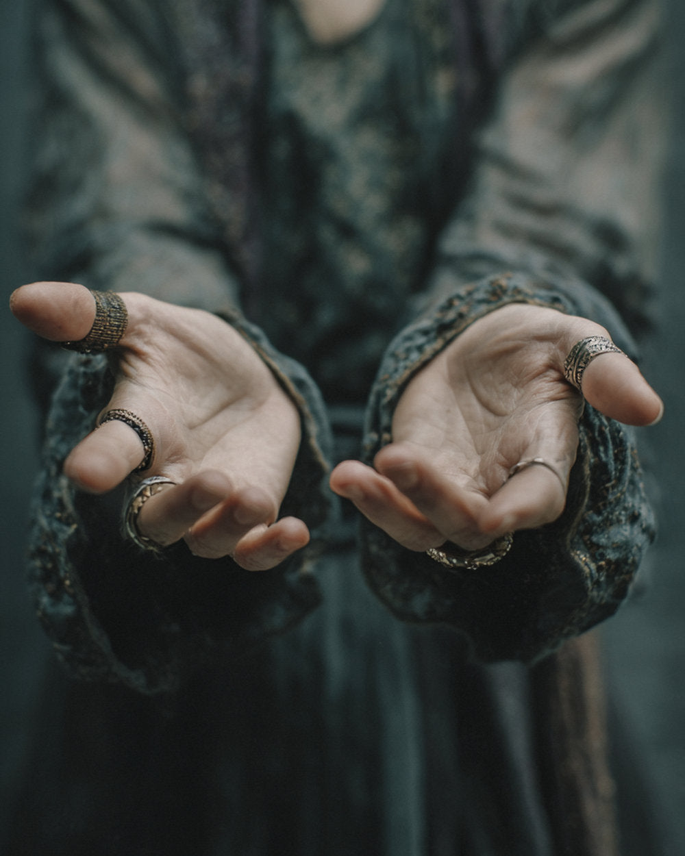Outstretched hands wearing silver rings, framed by dark embroidered clothing.