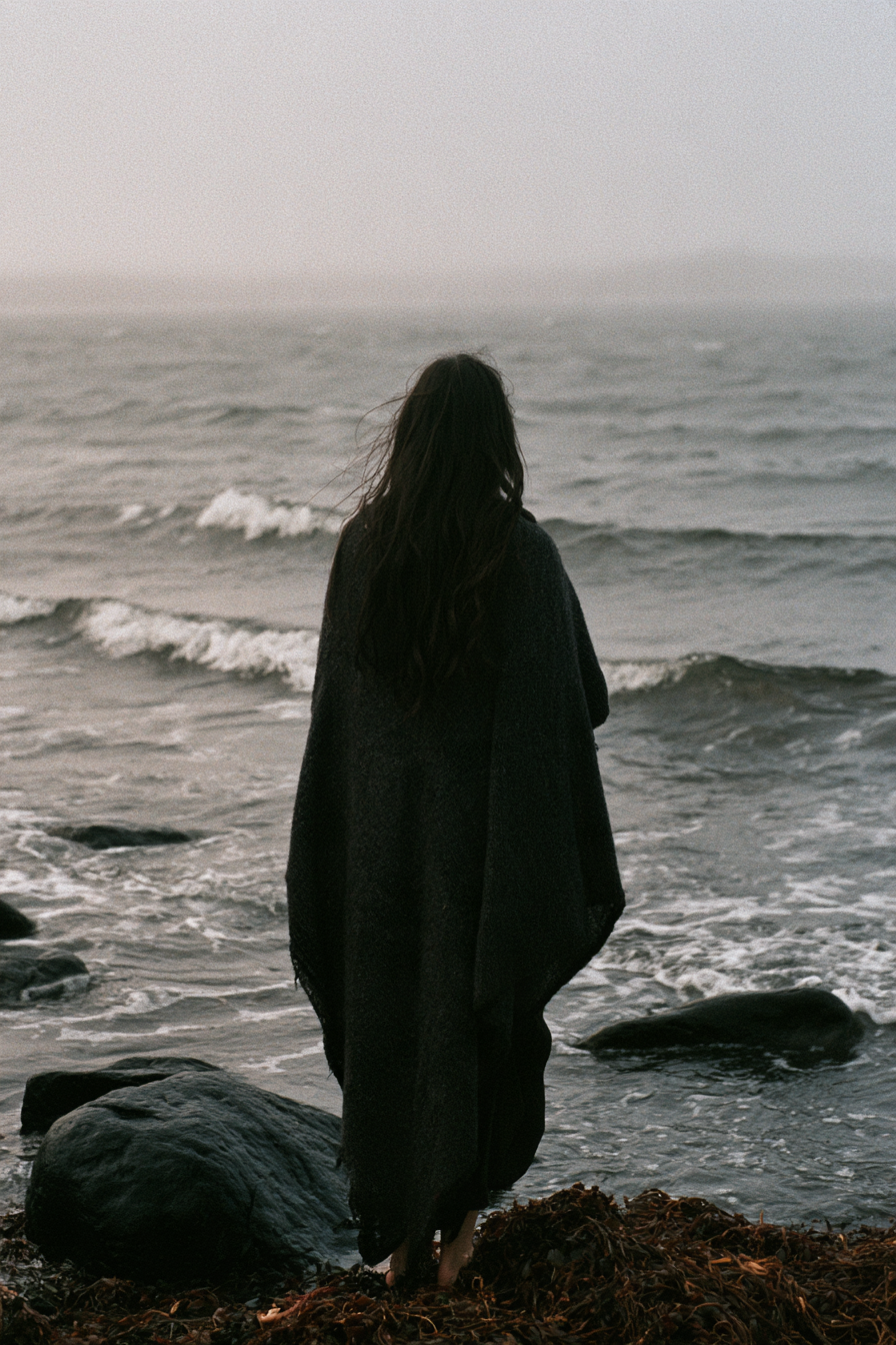 A woman in a dark wool shawl standing on black stones with her back to us, facing a grey northern sea.